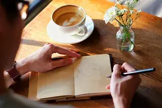 Person writing in a journal next to a coffee cup and flowers, symbolizing reflection and new beginnings after mission disruption