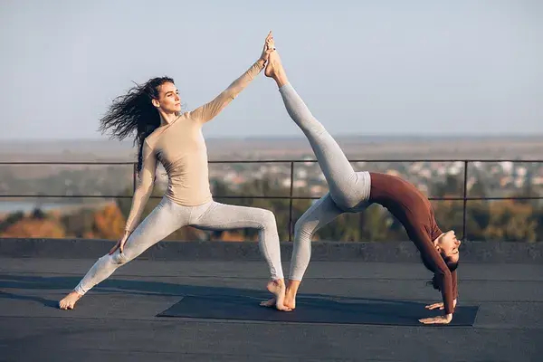 Two women performing an acro yoga pose on a rooftop with a scenic landscape in the background.