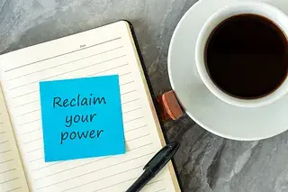 A blue sticky note on a notebook with the message “Reclaim your power”, next to a black pen and a cup of coffee on a gray table