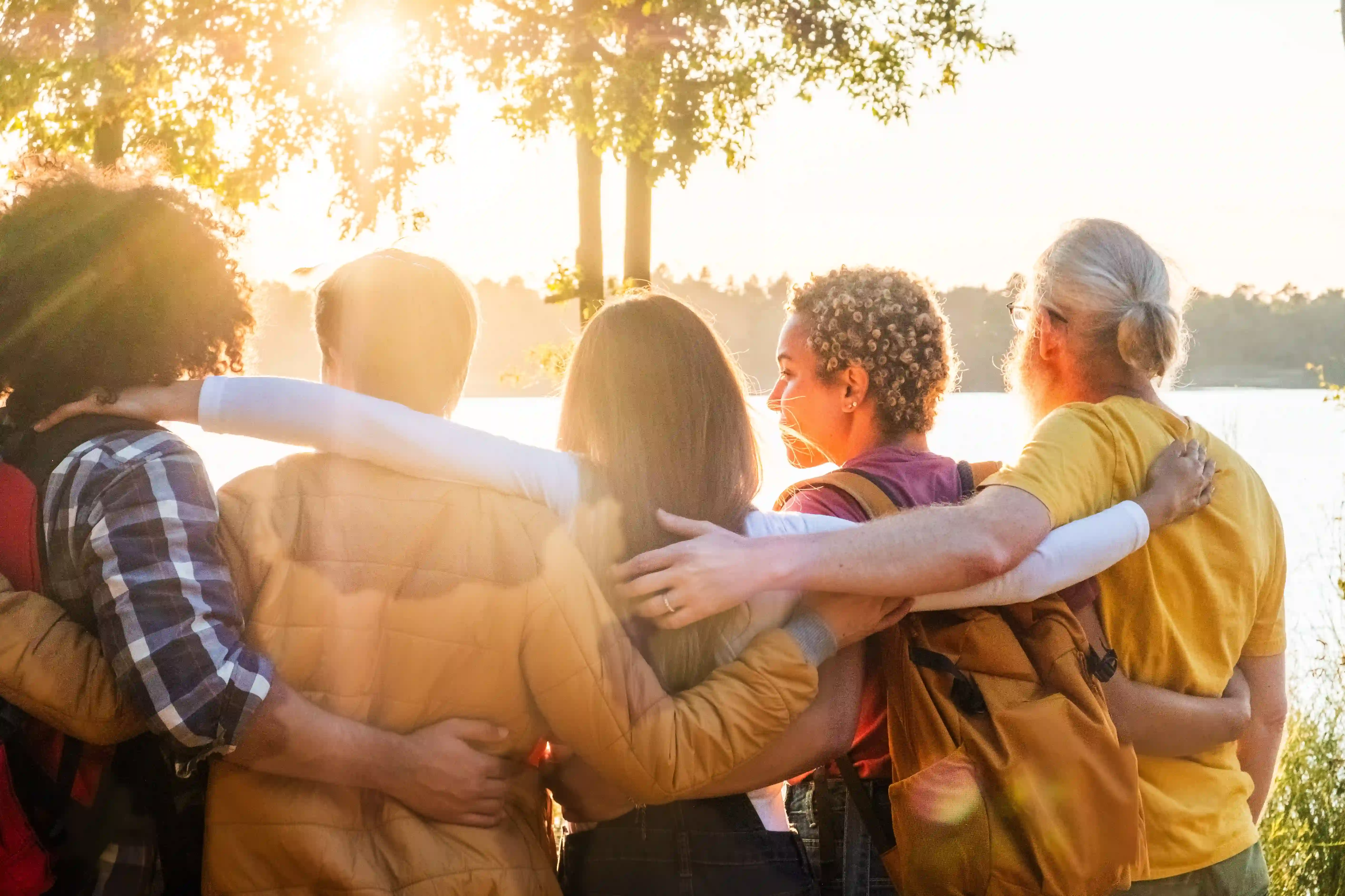 A diverse group of people standing together with arms around each other, facing the sunrise — representing unity, resilience, and staying human during the Trump era.